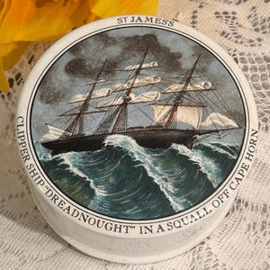 May include: A round white ceramic container lid featuring a detailed illustration of a clipper ship sailing through dark, turbulent ocean waves. The text ST JAMES'S and CLIPPER SHIP "DREADNOUGHT" IN A SQUALL OFF CAPE HORN is printed around the edge.