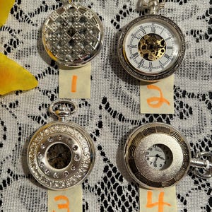 May include: Four silver pocket watches with different designs. One has a geometric pattern, another features Roman numerals and a visible mechanism, and two others have decorative details. Each watch is on a lace surface.