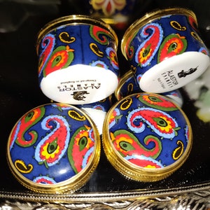 May include: A collection of small, round, blue enamel boxes with gold rims. Each box features a paisley design in red, yellow, and green. The text "ALASTOR ENAMELS" and "Designed in England" is visible on the base of the boxes.