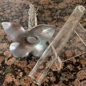 May include: A silver-colored, flower-shaped candle holder with a leaf-like accent and a clear glass tube resting on a clear plastic stand. The candle holder has a circular indentation in the center.