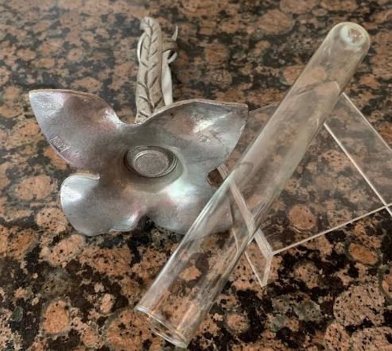 May include: A silver-colored, flower-shaped candle holder with a leaf-like accent and a clear glass tube resting on a clear plastic stand. The candle holder has a circular indentation in the center.