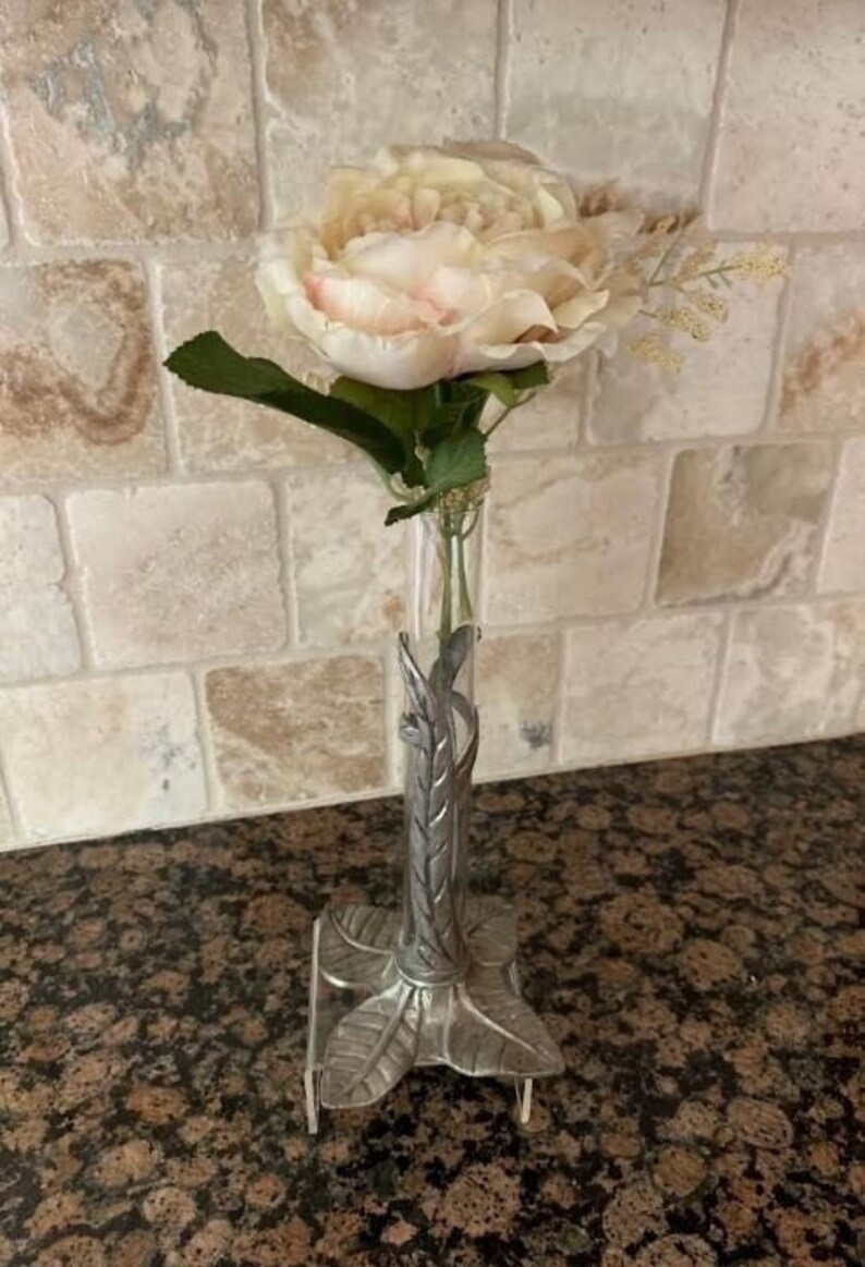 May include: A clear glass bud vase with a silver-colored metal base and accents, holding a light pink and cream-colored flower with green leaves. The vase has a decorative leaf design.