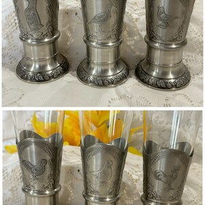 Op de afbeelding: Set van zes heldere pilsglazen met tinnen bases. Elke basis heeft een ander vogelontwerp, waaronder een eend, fazant en haan, omringd door tarwe. De glazen zijn geschikt voor het serveren van bier of andere dranken.