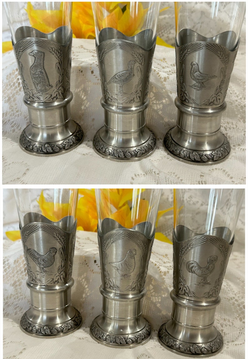 Op de afbeelding: Set van zes heldere pilsglazen met tinnen bases. Elke basis heeft een ander vogelontwerp, waaronder een eend, fazant en haan, omringd door tarwe. De glazen zijn geschikt voor het serveren van bier of andere dranken.