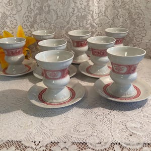May include: A set of eight white porcelain teacups with matching saucers. Each cup features a red band with a decorative design and text. The saucers have a red border. The cups are arranged on a white lace tablecloth.
