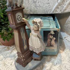 May include: A porcelain figurine depicting a young girl reaching towards a grandfather clock, with a cat perched on top. The clock is brown and beige, and the girl wears a white apron and bonnet. The figurine is on a gray base.