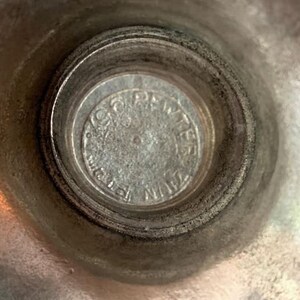 May include: Close-up of a silver-colored metal object. The center features a circular emblem with embossed text. The surrounding area has a textured, slightly tarnished appearance, suggesting age or use. The object's design is simple and functional.
