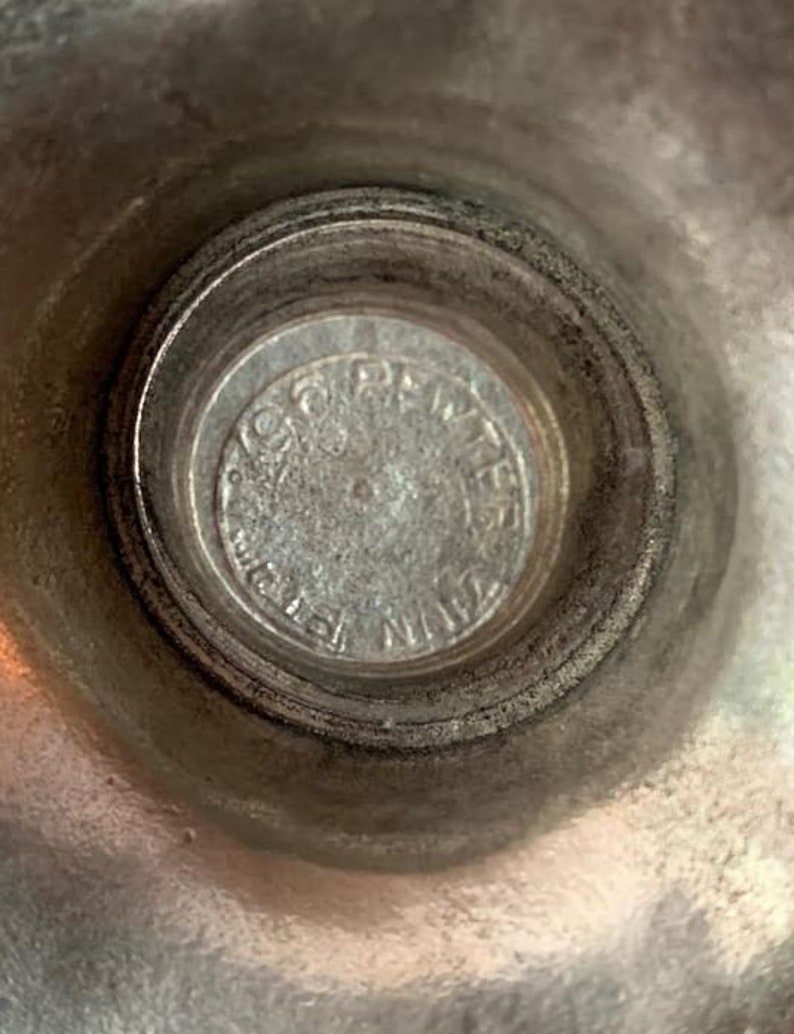 May include: Close-up of a silver-colored metal object. The center features a circular emblem with embossed text. The surrounding area has a textured, slightly tarnished appearance, suggesting age or use. The object's design is simple and functional.