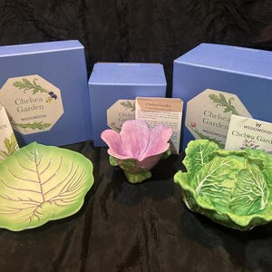 May include: A collection of Wedgwood Chelsea Garden ceramic pieces, including a leaf-shaped dish, a cabbage-shaped bowl, and a pink flower-shaped bowl. The items are accompanied by blue boxes and informational cards.