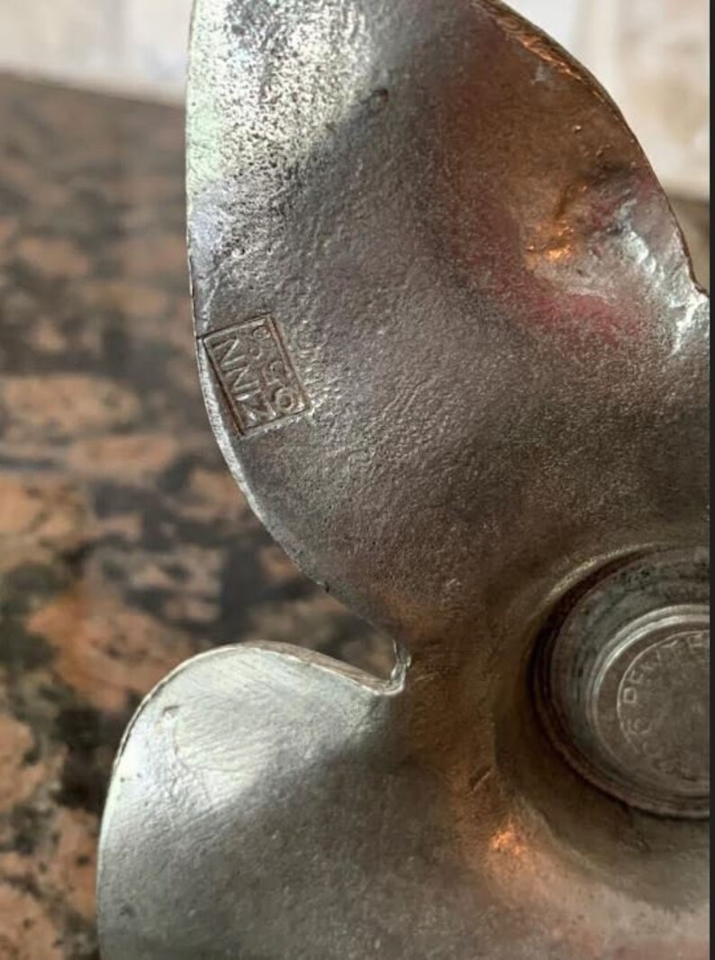 May include: Close-up of a vintage metal object with a textured, silver-gray finish. The object has a curved, wing-like shape with a circular element on the right. The text "ZINN 0.50" is visible. The object appears to be a decorative or functional piece.