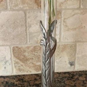 May include: A clear glass bud vase with a silver-colored metal leaf design. The vase holds greenery and small white flowers. The base of the vase has a leaf design. The vase is approximately 10 inches tall.