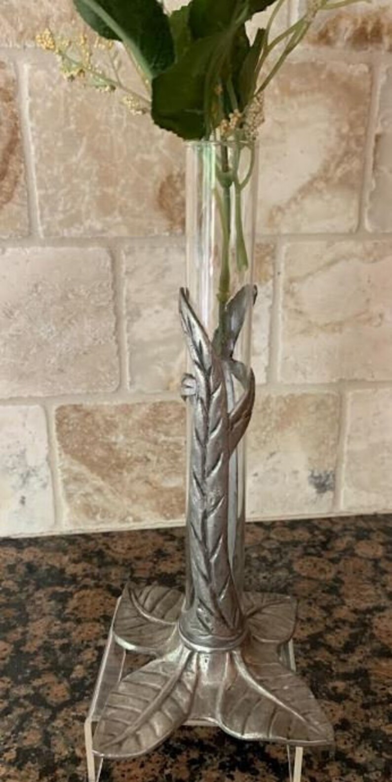 May include: A clear glass bud vase with a silver-colored metal leaf design. The vase holds greenery and small white flowers. The base of the vase has a leaf design. The vase is approximately 10 inches tall.