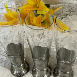 Op de afbeelding: Drie heldere bierglazen met tinnen voetjes. De voetjes hebben decoratieve ge&euml;tste ontwerpen. Gele bloemen staan op de achtergrond. De glazen staan op een wit kanten tafelkleed.