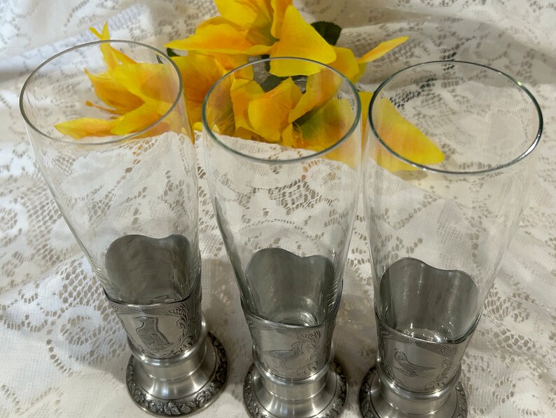 Op de afbeelding: Drie heldere bierglazen met tinnen voetjes. De voetjes hebben decoratieve ge&euml;tste ontwerpen. Gele bloemen staan op de achtergrond. De glazen staan op een wit kanten tafelkleed.