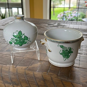 May include: Two white ceramic pieces with green floral designs and gold trim. One is a round vase, the other a small handled pot. Both pieces have a classic, elegant aesthetic, suitable for home decor.