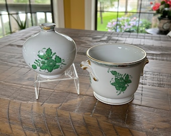 Op de afbeelding: Twee witte keramische stukken met groene bloemmotieven en gouden randen. Eén is een ronde vaas, de andere een kleine pot met handvatten. Beide stukken hebben een klassieke, elegante uitstraling, geschikt voor woondecoratie.