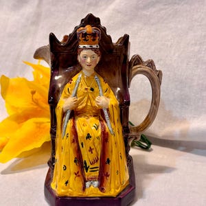 May include: A vintage ceramic pitcher featuring a regal figure in a yellow gown and crown, seated on a brown throne. The pitcher has a handle and spout, with a dark purple base and accents. The overall design is ornate and detailed.