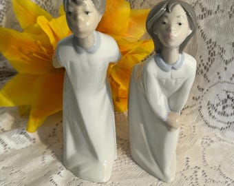 Op de afbeelding: Twee porseleinen beeldjes van kinderen, één staand en één voorovergebogen. Beide figuren dragen witte gewaden met lichtblauwe kragen. De beeldjes hebben grijs haar en staan tegen een gele bloem en een kanten achtergrond.