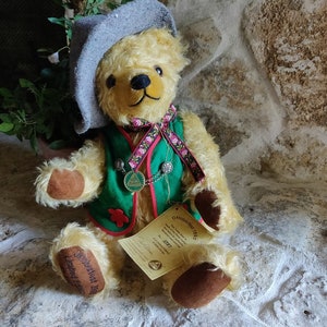 May include: A plush teddy bear wearing a green vest with red trim and a gray hat. The bear has a ribbon with pink roses and a small metal pendant. The bear's paws are brown, and a tag reads "Oktoberfest 1995".