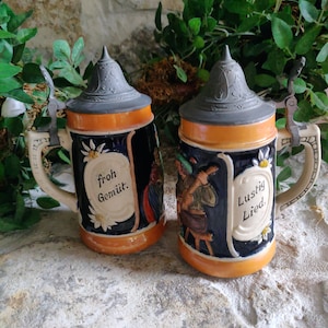 May include: Two ceramic beer steins with pewter lids. The steins have a black base with orange trim and cream-colored handles. One stein reads "froh Gemüt" and the other reads "Lustig Lied."