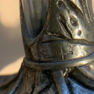May include: Close-up of a pewter-colored decorative object. The object has a detailed, textured surface with leaf-like patterns and a cylindrical shape. The design appears to be vintage or antique.