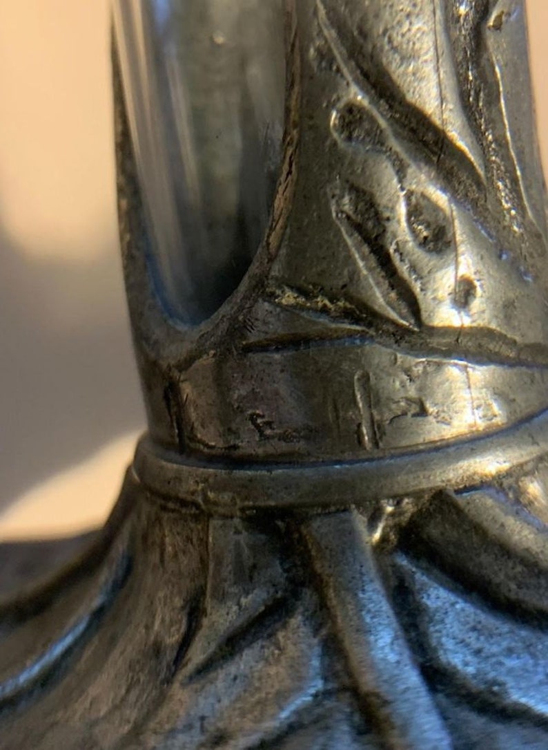 May include: Close-up of a pewter-colored decorative object. The object has a detailed, textured surface with leaf-like patterns and a cylindrical shape. The design appears to be vintage or antique.