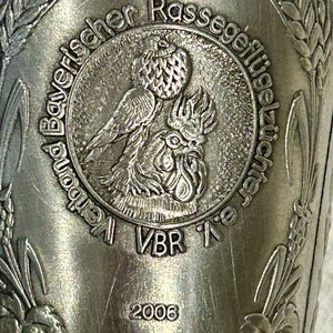 Op de afbeelding: Close-up van een zilveren metalen beker met reli&euml;fdetails. De beker heeft een centraal embleem met een vogel- en haanontwerp, omringd door tekst. Het jaar 2006 is zichtbaar onder het embleem, samen met extra tekst en tarwearen.