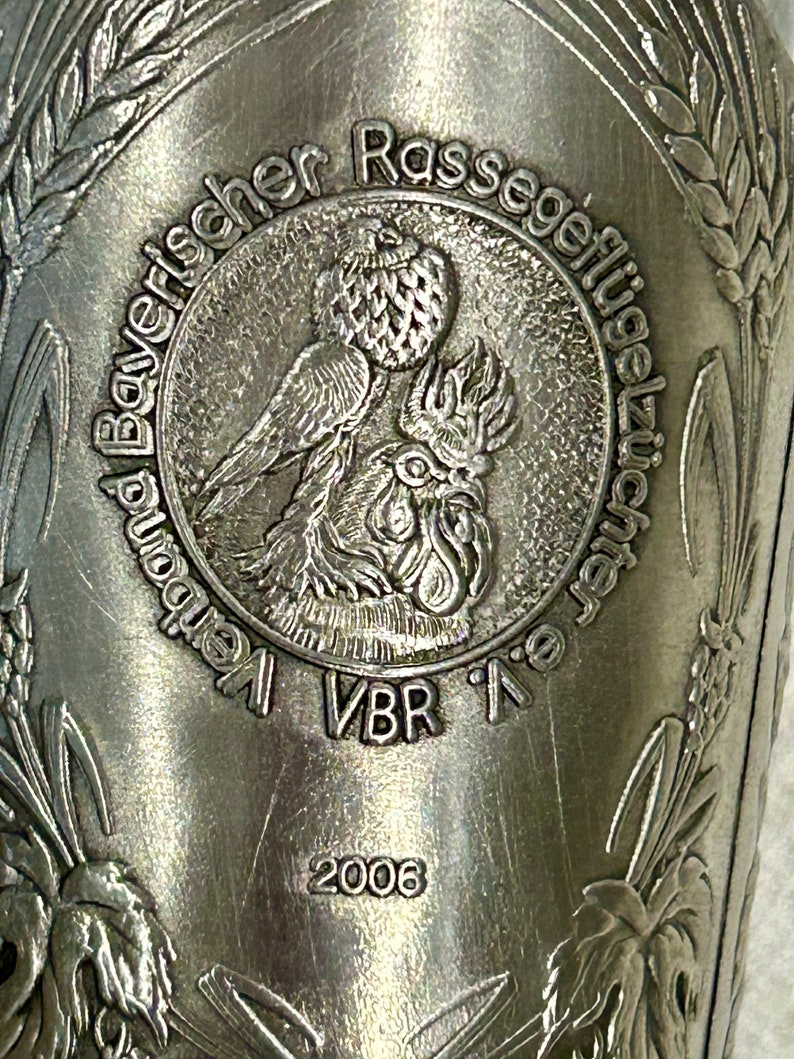 Op de afbeelding: Close-up van een zilveren metalen beker met reli&euml;fdetails. De beker heeft een centraal embleem met een vogel- en haanontwerp, omringd door tekst. Het jaar 2006 is zichtbaar onder het embleem, samen met extra tekst en tarwearen.