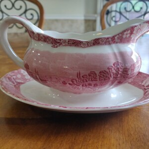 May include: A white and pink gravy boat with a matching saucer. The boat has a curved handle and a scalloped rim. The design features a landscape scene and floral accents. The set is made of ceramic.