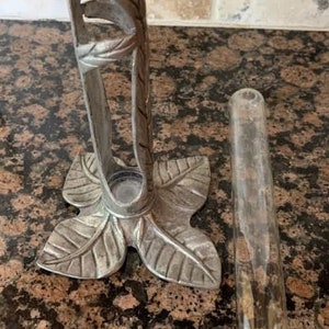 May include: A silver-colored metal bud vase holder with a leaf design base and a clear glass tube. The holder has a decorative leaf and stem design. The vase is designed to hold a single flower stem.