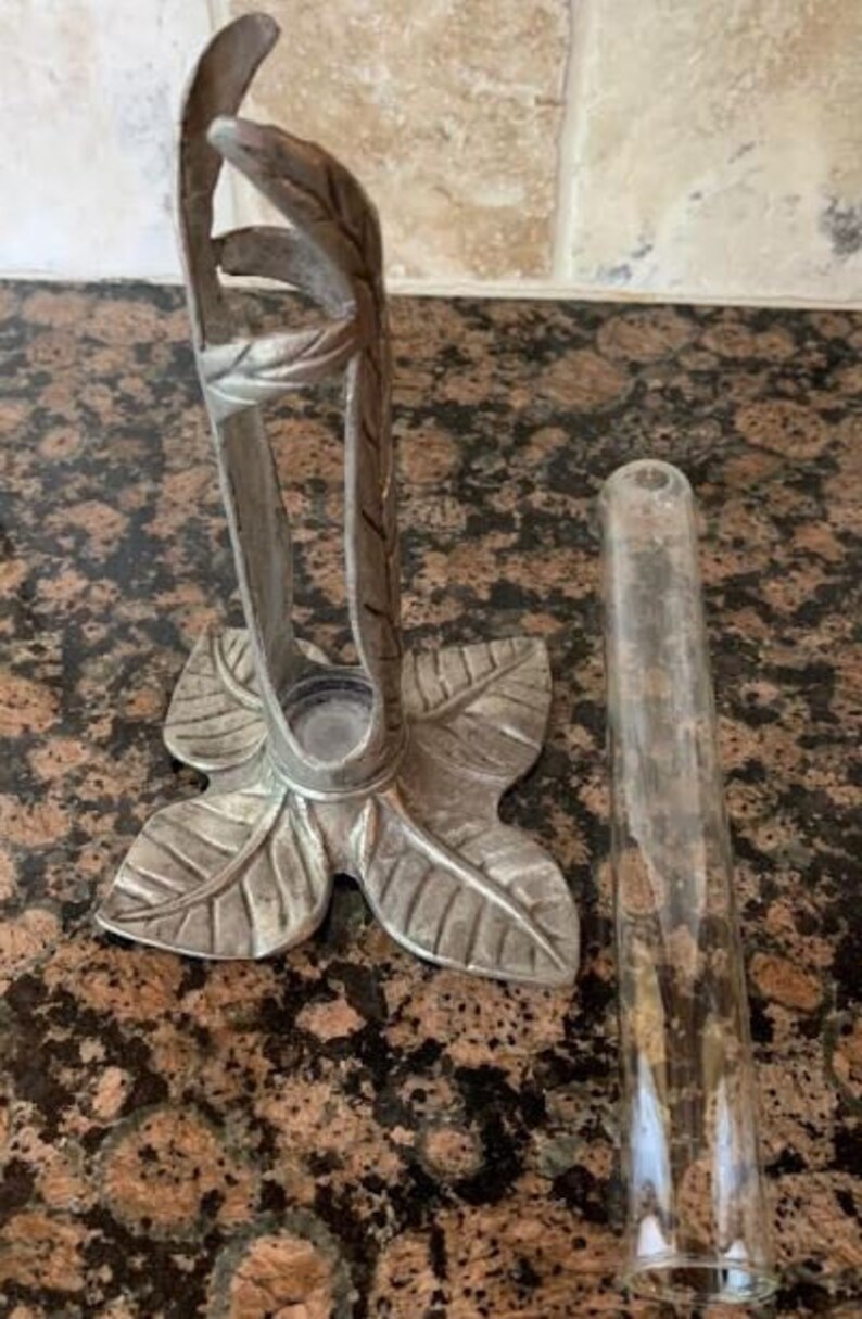 May include: A silver-colored metal bud vase holder with a leaf design base and a clear glass tube. The holder has a decorative leaf and stem design. The vase is designed to hold a single flower stem.