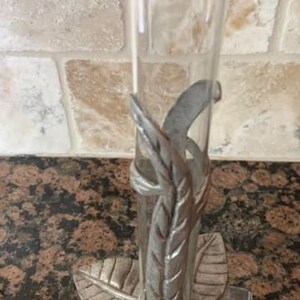 May include: Clear glass bud vase with a decorative metal leaf design. The vase is supported by a metal base shaped like a flower with three leaves. The metal has a silver finish. The vase is approximately 8 inches tall.