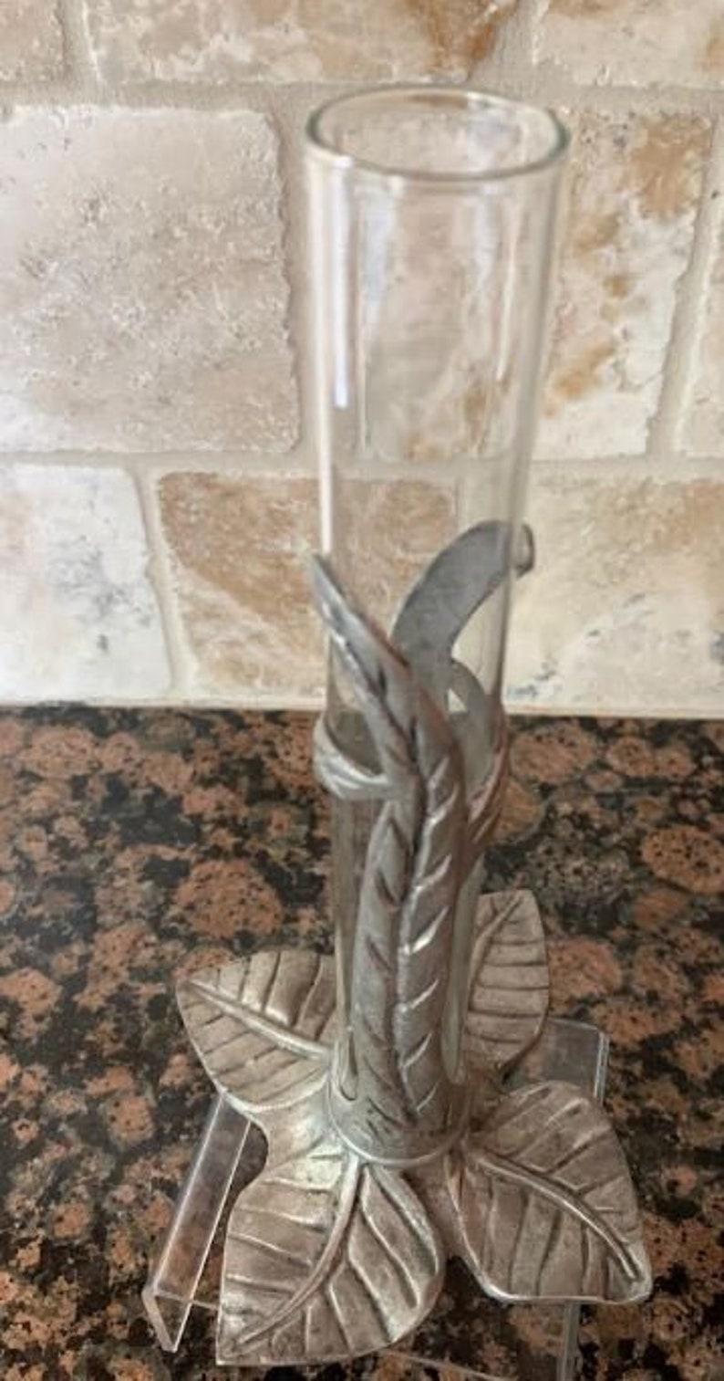 May include: Clear glass bud vase with a decorative metal leaf design. The vase is supported by a metal base shaped like a flower with three leaves. The metal has a silver finish. The vase is approximately 8 inches tall.