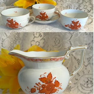 May include: White porcelain teacups and a creamer with orange floral designs. The teacups have gold trim and handles. The creamer has a curved handle and a decorative rim. The set is displayed on a white lace tablecloth.