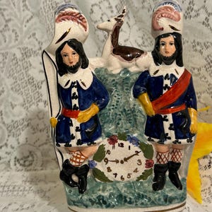 May include: A vintage ceramic decorative piece featuring two figures in blue coats and hats, flanking a clock and a deer. The figures have black hair and beards. The clock is surrounded by flowers. The piece has a crackled finish.