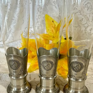 Op de afbeelding: Drie heldere bierpilsnerglazen met tinnen bases en decoratieve accenten. Elk glas heeft een gedetailleerde tinnen basis met een cirkelvormig embleem en tarwe- en druivenontwerpen. De glazen worden gepresenteerd tegen een achtergrond van gele bloemen.