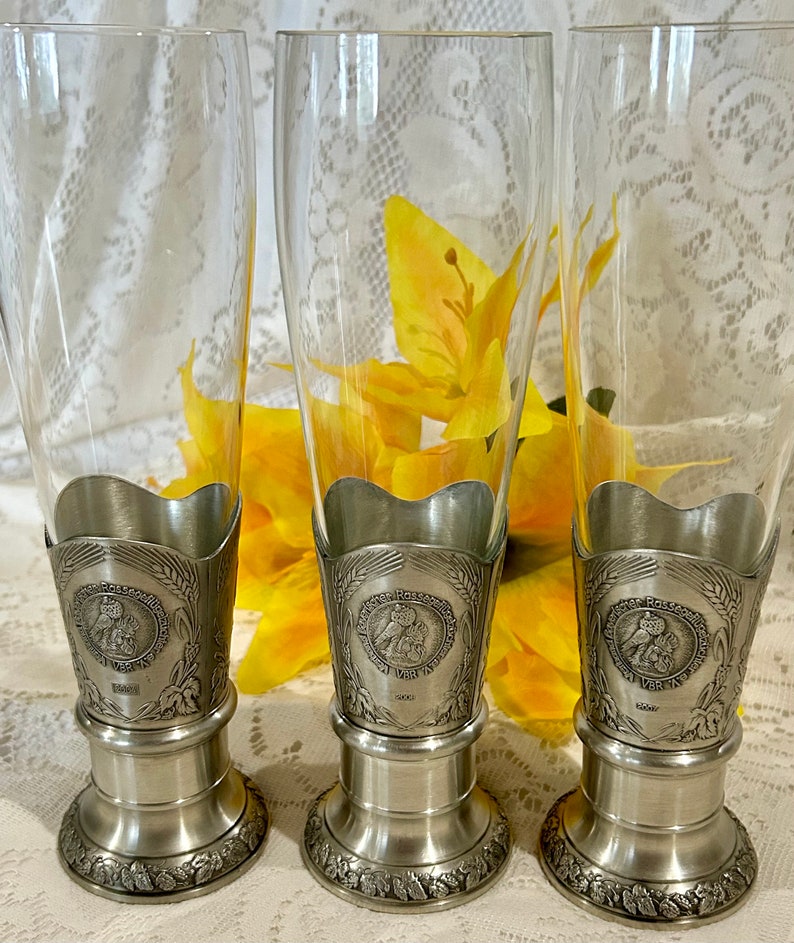 Op de afbeelding: Drie heldere bierpilsnerglazen met tinnen bases en decoratieve accenten. Elk glas heeft een gedetailleerde tinnen basis met een cirkelvormig embleem en tarwe- en druivenontwerpen. De glazen worden gepresenteerd tegen een achtergrond van gele bloemen.
