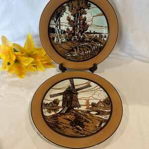 May include: Two decorative plates with a brown border and landscape scenes. One plate features trees and a body of water, while the other depicts a windmill. The plates have a black trim and are set against a white background.