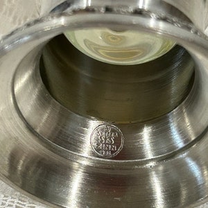 Op de afbeelding: Close-up van een zilverkleurige metalen kandelaar met een brede, uitlopende basis en een centrale opening. Een heldere glazen bol is van binnen zichtbaar. De basis heeft een cirkelvormig embleem met tekst. De kandelaar is waarschijnlijk vintage.