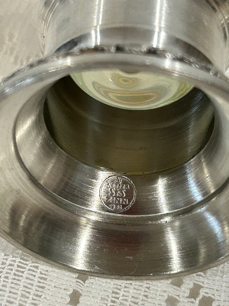 Op de afbeelding: Close-up van een zilverkleurige metalen kandelaar met een brede, uitlopende basis en een centrale opening. Een heldere glazen bol is van binnen zichtbaar. De basis heeft een cirkelvormig embleem met tekst. De kandelaar is waarschijnlijk vintage.