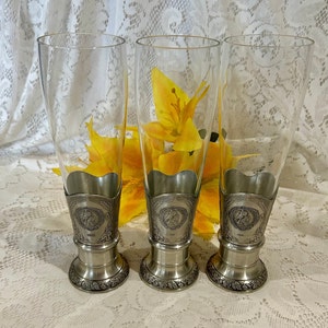 Op de afbeelding: Drie heldere pilsglazen met zilverkleurige metalen bases en decoratieve banden. De bases hebben een bloemenontwerp en de banden hebben een cirkelvormig embleem. Gele bloemen zijn zichtbaar achter de glazen.