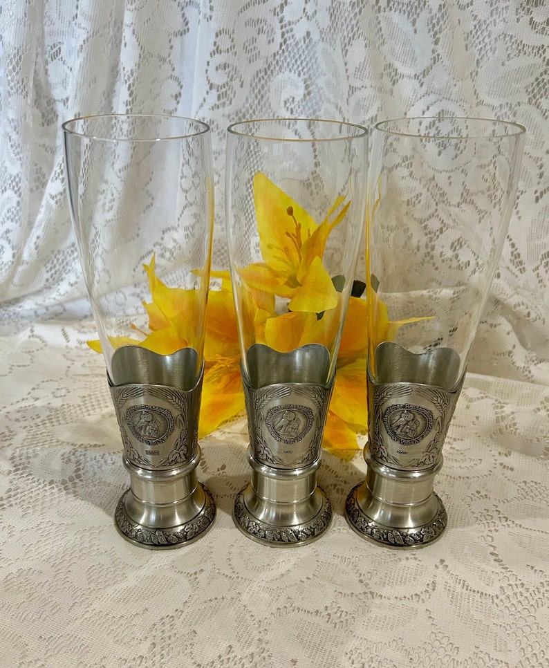Op de afbeelding: Drie heldere pilsglazen met zilverkleurige metalen bases en decoratieve banden. De bases hebben een bloemenontwerp en de banden hebben een cirkelvormig embleem. Gele bloemen zijn zichtbaar achter de glazen.