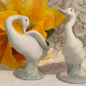 May include: Two white ceramic goose figurines. One goose has its head turned back, and the other has its head up. The figurines have a light blue base with green and tan accents. The background includes a large yellow flower.