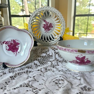 May include: A white porcelain teacup saucer, bowl, and decorative basket with a floral design in fuchsia and gold trim. The teacup saucer has a scalloped edge. The bowl sits on a pedestal base.
