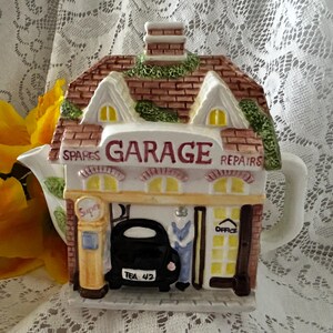 May include: A ceramic teapot shaped like a garage, with the words "GARAGE" and "REPAIRS" on the front. The teapot features a brick roof, a chimney, and a small black car. The words "SPARES" and "OFFICE" are also visible.