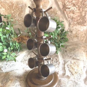 May include: A wooden mug tree with a round base and a central column supporting multiple pegs. Eight small, silver-colored metal mugs are suspended from the pegs. The tree is made of dark brown wood and has a decorative top.