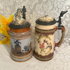 May include: Two ceramic beer steins with hinged metal lids. The left stein features a blue-clad figure on the lid and a scene of figures working with barrels. The right stein has a textured lid and a scene of two figures dancing with mugs.
