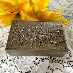May include: A rectangular metal box with a detailed relief scene of soldiers in historical attire. The box is silver-toned and sits on a white lace surface, with a yellow flower in the background. The box's design features soldiers with spears and swords.