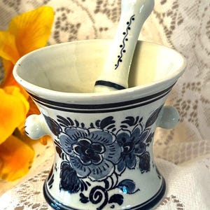 May include: A white ceramic mortar and pestle set. The mortar features blue floral designs and a dark blue rim. The pestle is white with a dark blue band and decorative accents. The set includes two small handles on the sides of the mortar.