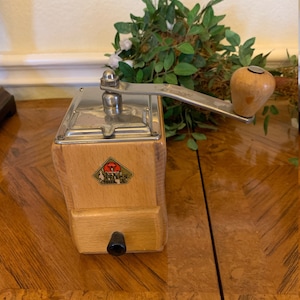 May include: A vintage coffee grinder with a wooden body and a metal top and handle. The grinder has a small drawer and a logo on the front. The handle is made of wood. The grinder is sitting on a wooden surface.